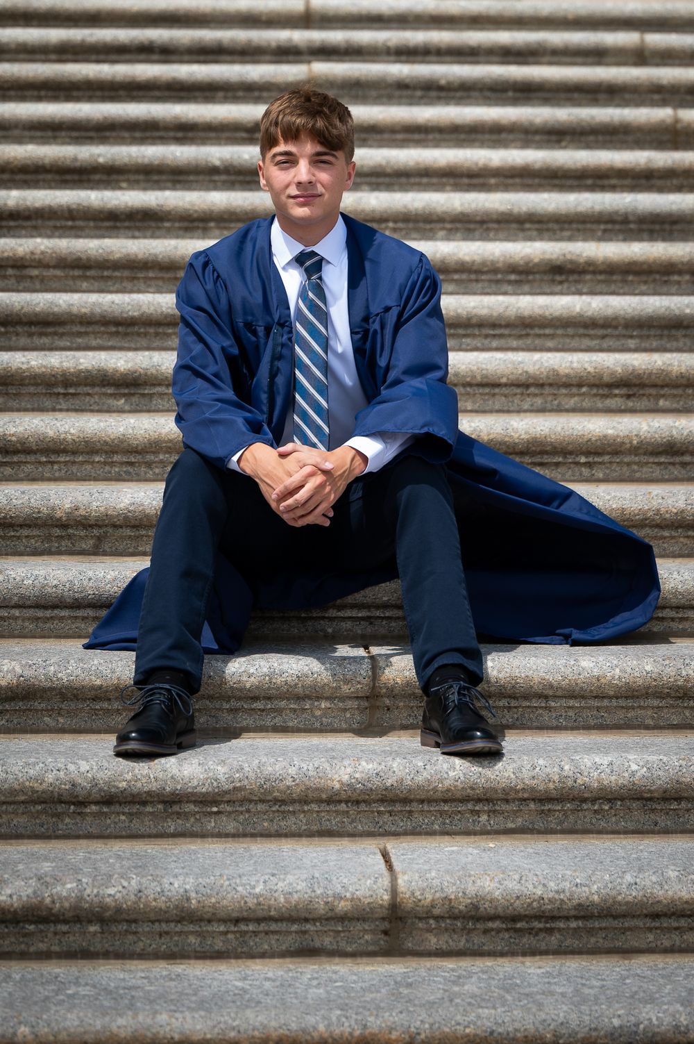 Young man in blue graduation gown and tie sitting on stone steps, smiling with hands clasped.