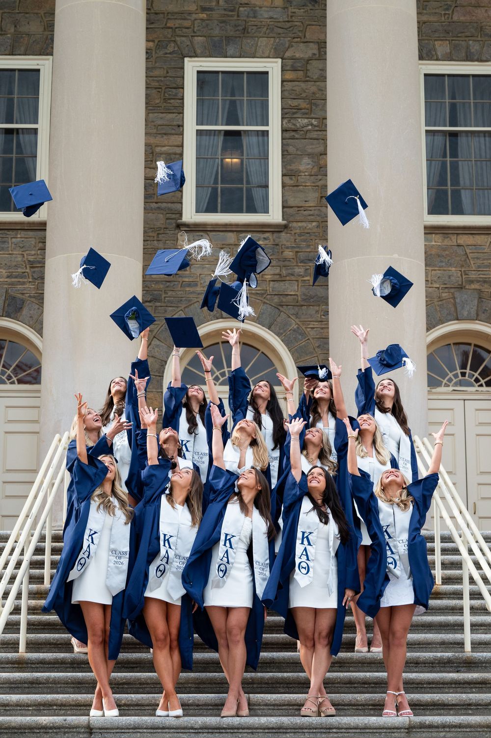 Group of female graduates in navy robes tossing caps on university steps, wearing Kappa Alpha Theta sorority attire.