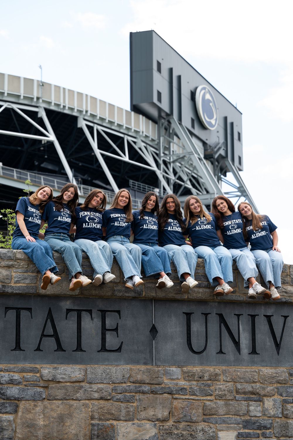 Eight women in matching Penn State Alumni shirts sit on a stone university sign in front of Beaver Stadium.