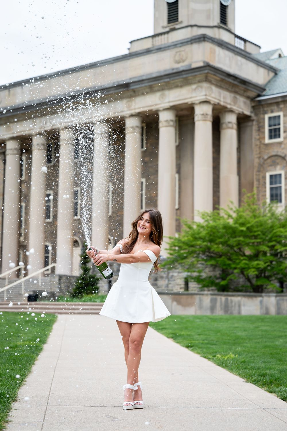 Woman in white dress popping champagne bottle in front of a classic stone building with columns.