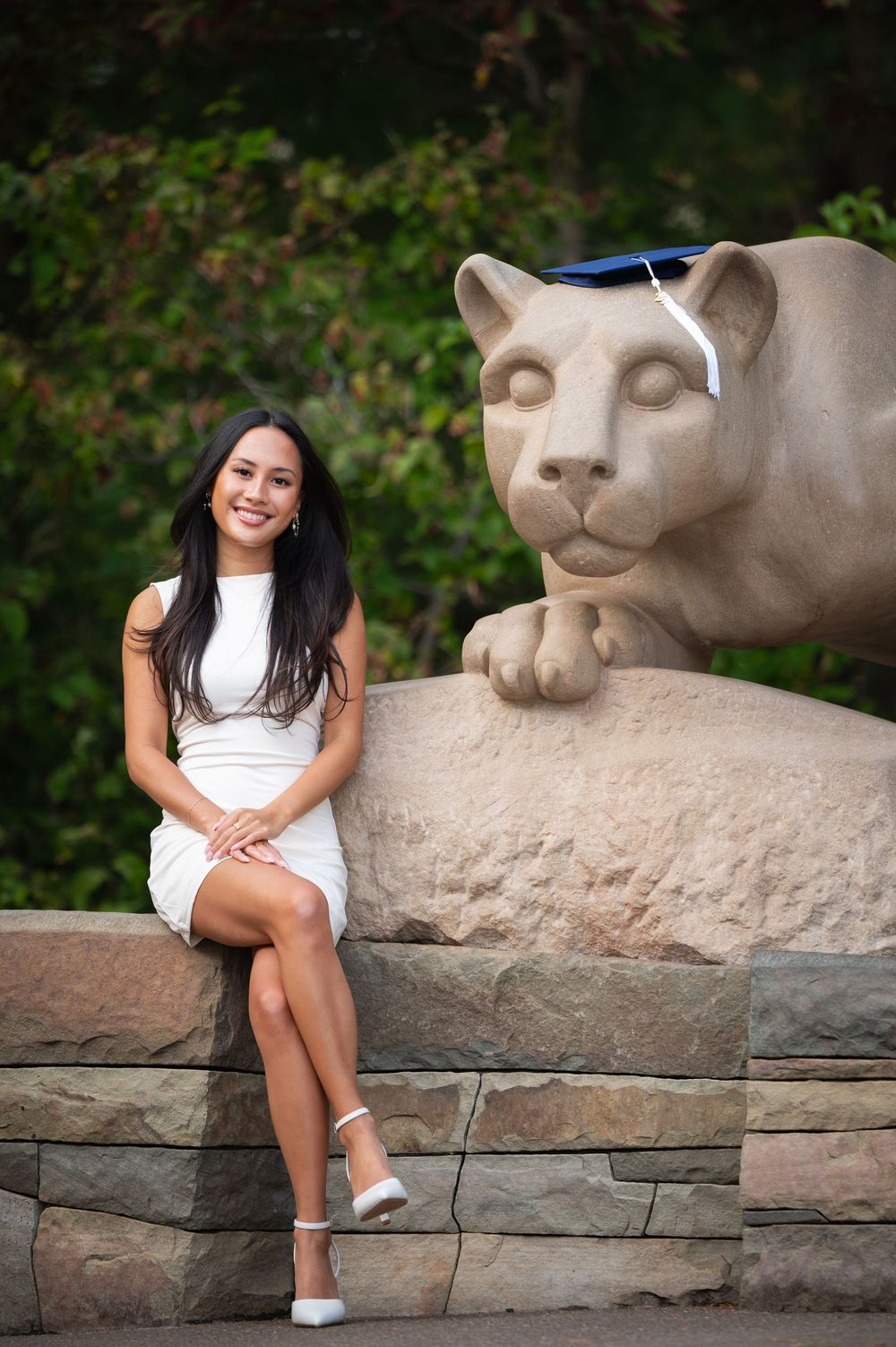 Student sits next to iconic Penn State Nittany Lion statue on campus stone wall during graduation photo session.