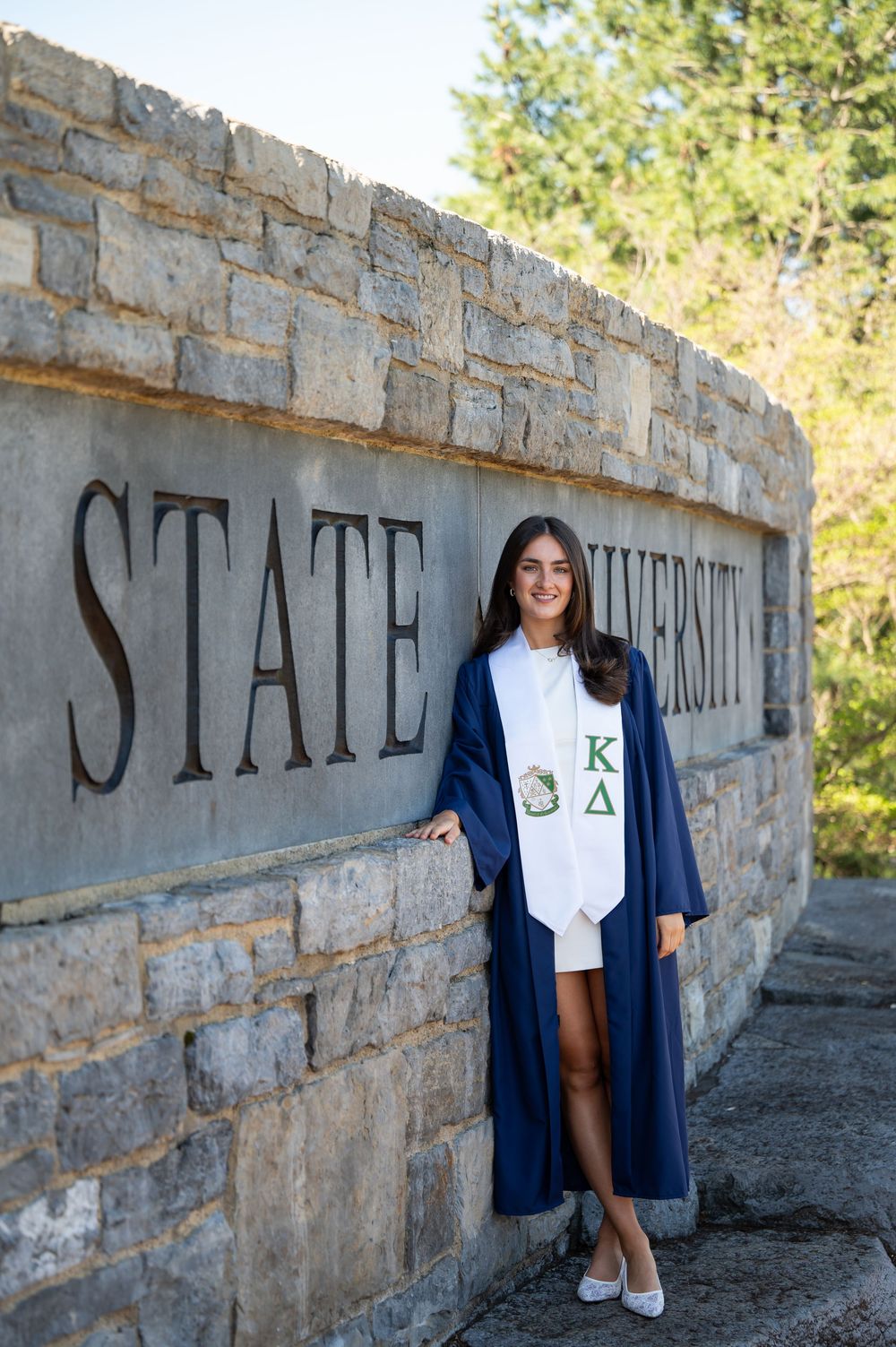 College graduate in navy cap and gown with Kappa Delta stole posing by a State University stone sign.