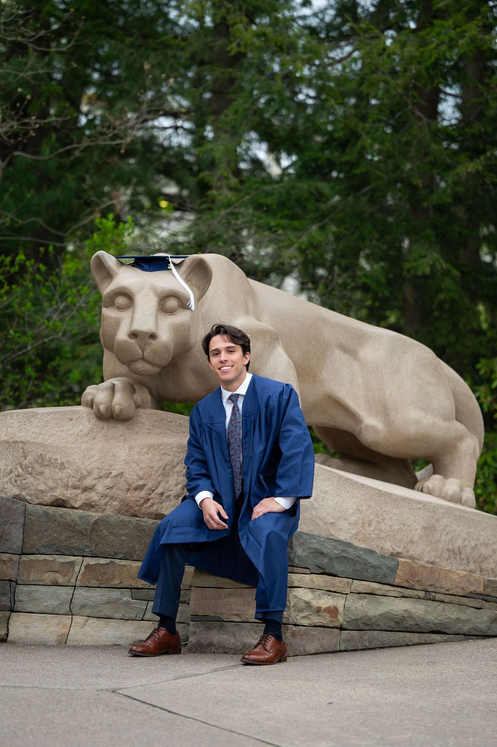Penn State graduate in blue gown sits proudly before the iconic Nittany Lion shrine during commencement celebration.