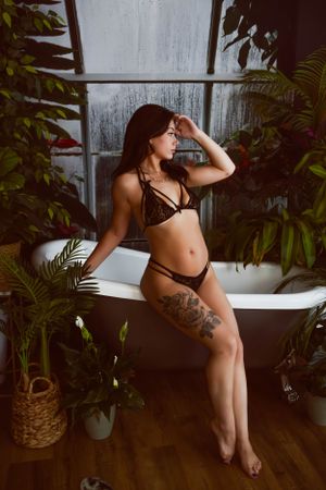 Person in brown lingerie poses in tropical bathroom setting with plants and bathtub against window.