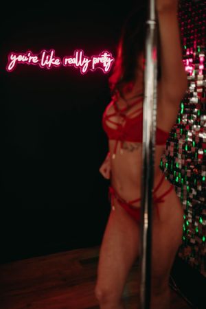 A dimly lit club scene with a neon pink sign reading 'you're like really pretty' above a pole and sequined backdrop.