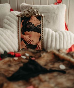 Artistic boudoir photograph of a mirror on white bedding reflecting black lace lingerie with scattered rose petals.