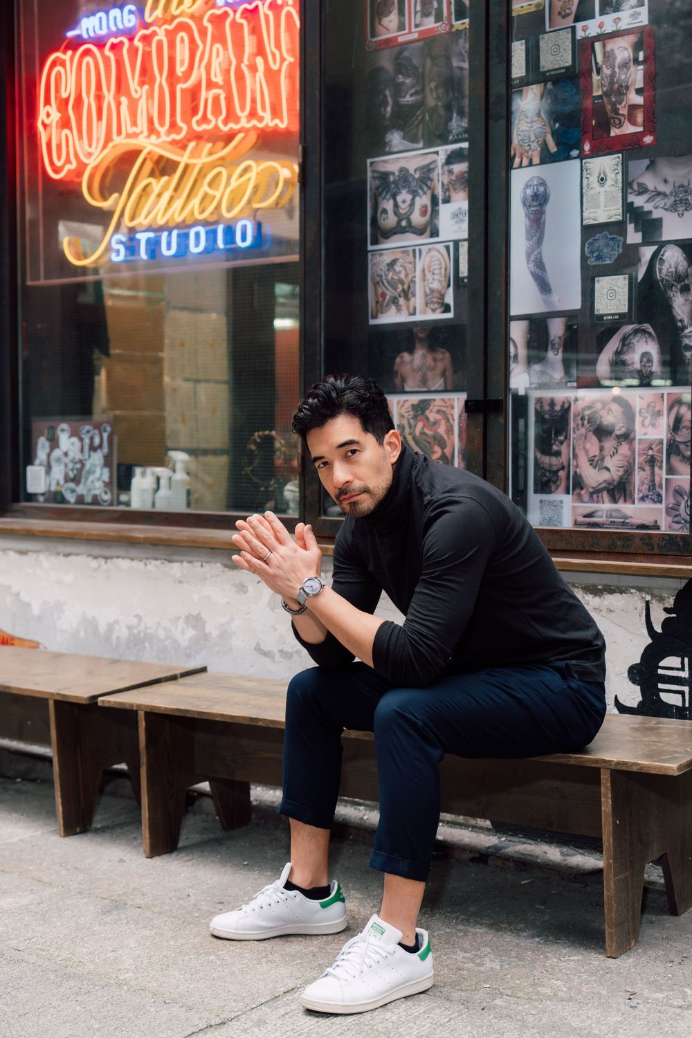 Person in black sweater and jeans sits on bench outside tattoo studio with neon sign and photo-covered window.