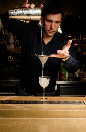 Bartender pouring cocktail from shaker into martini glass with precise technique.