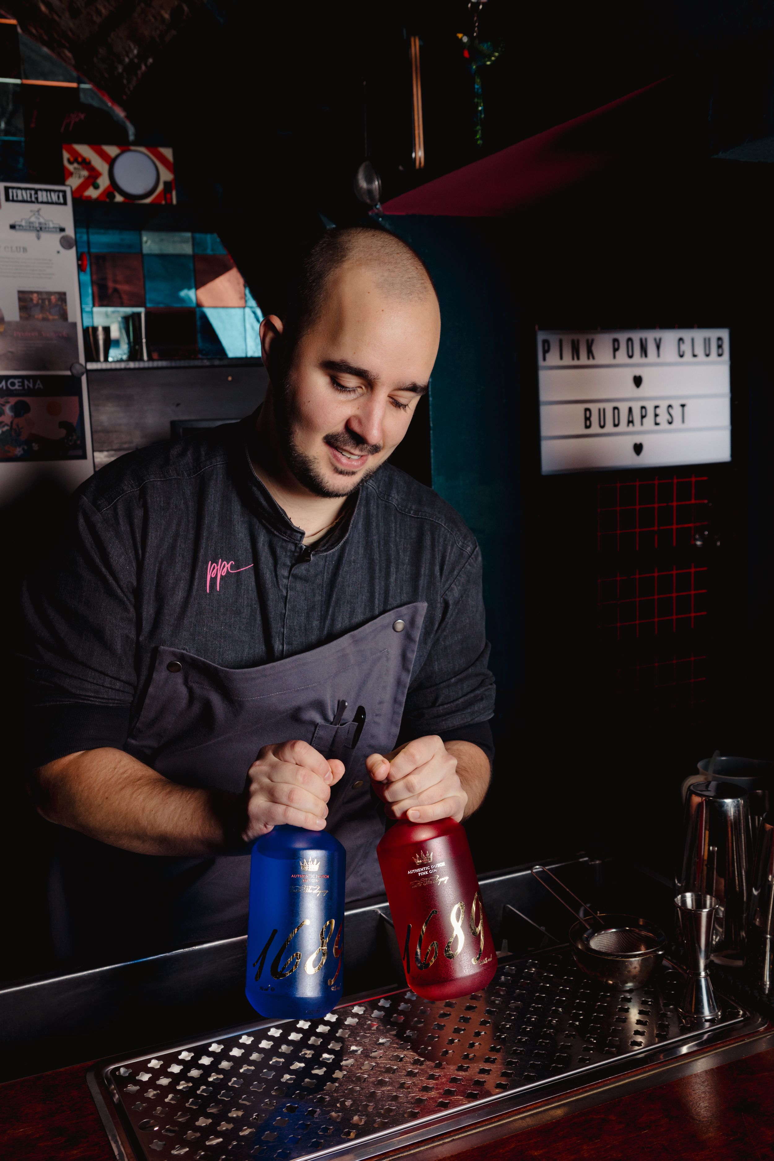 Bartender in dark apron manipulates red and blue cocktail shakers at dimly lit bar.