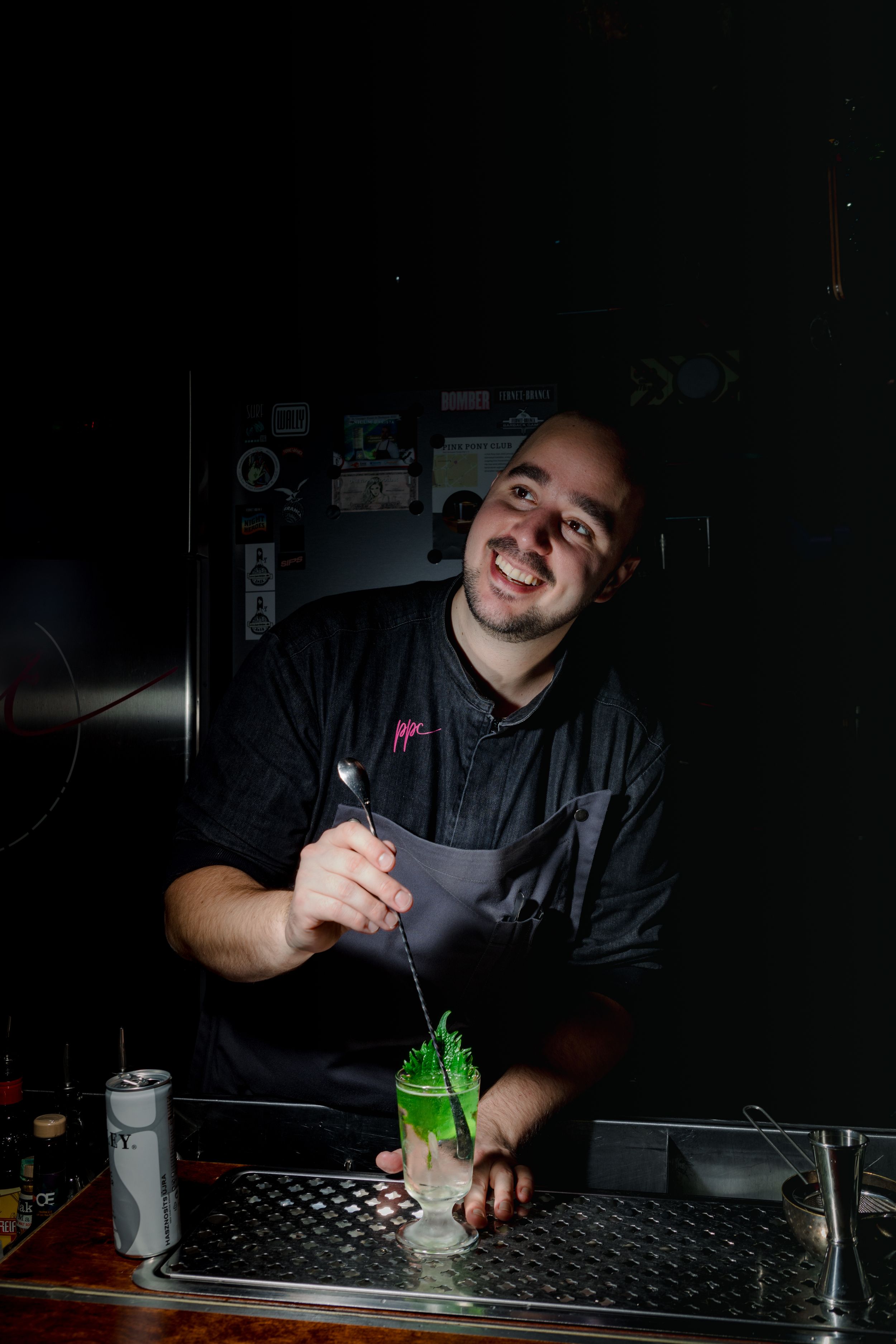 Bartender in dark apron pours bright green cocktail in moody bar lighting.