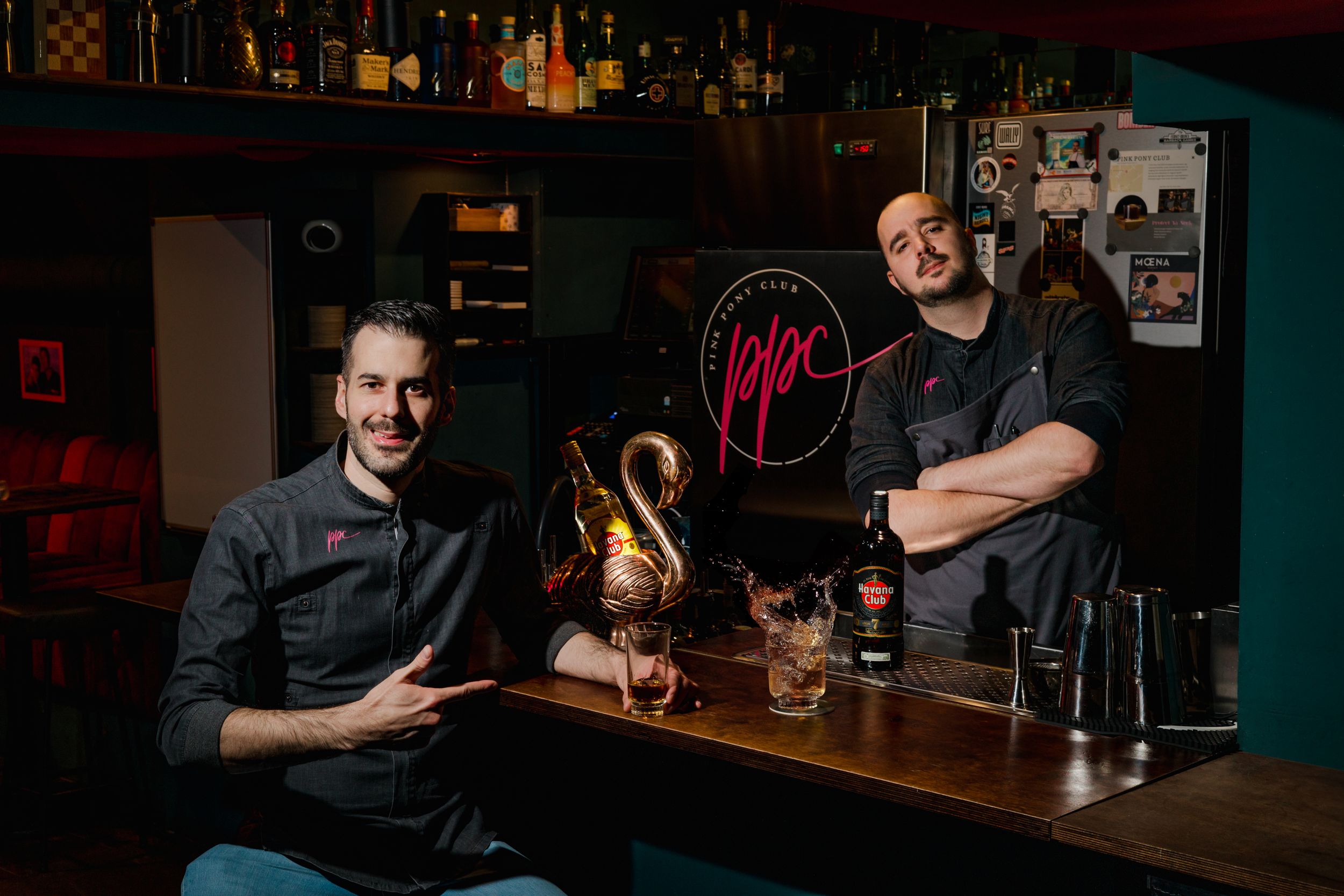 Bartenders behind counter with bottles and glassware in dark moody bar setting.