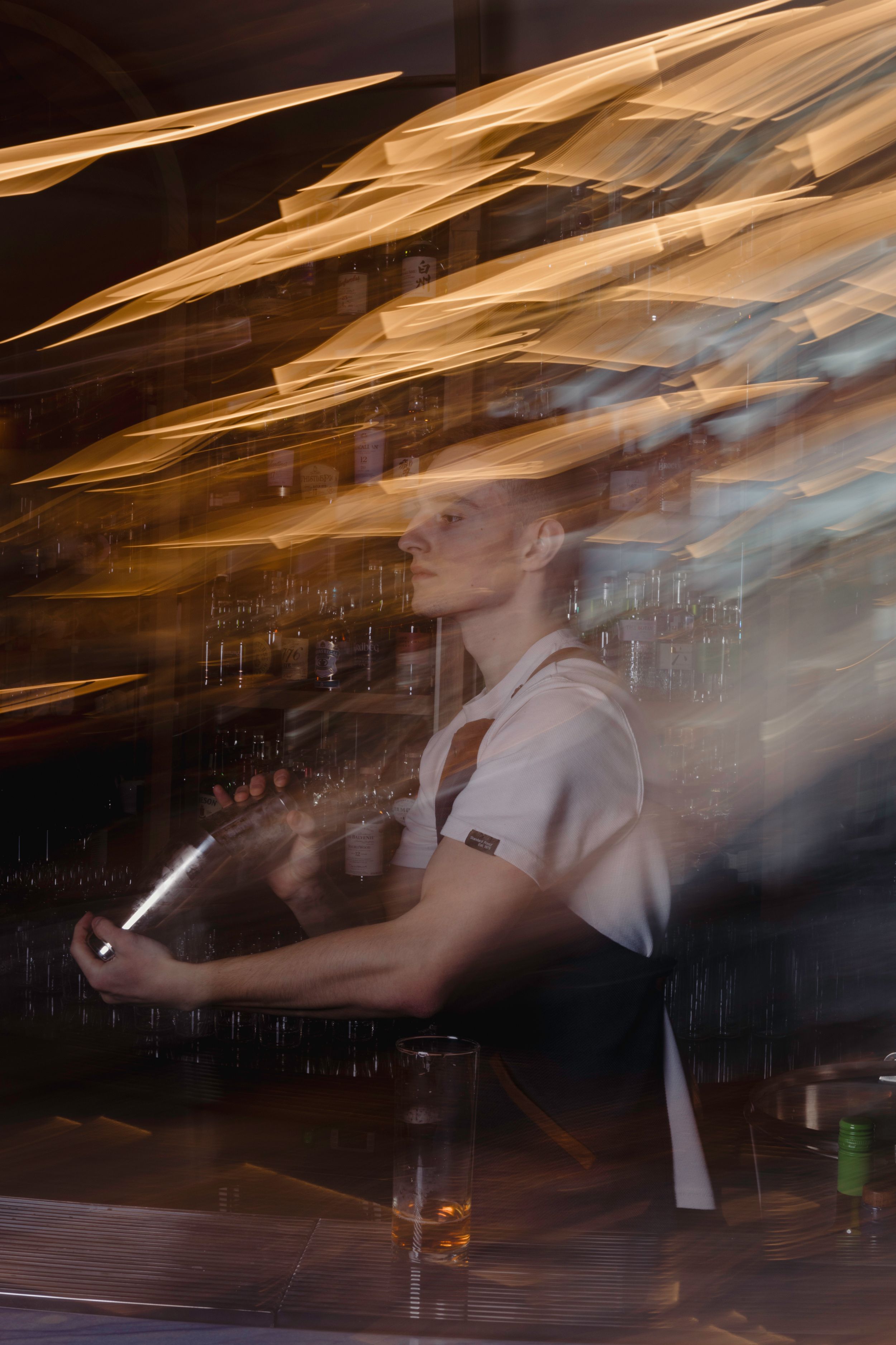 Motion blur capture of bartender mixing cocktails behind bar counter in artistic lighting.