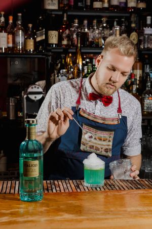 Bartender in apron and bow tie preparing green cocktail at wooden bar counter.