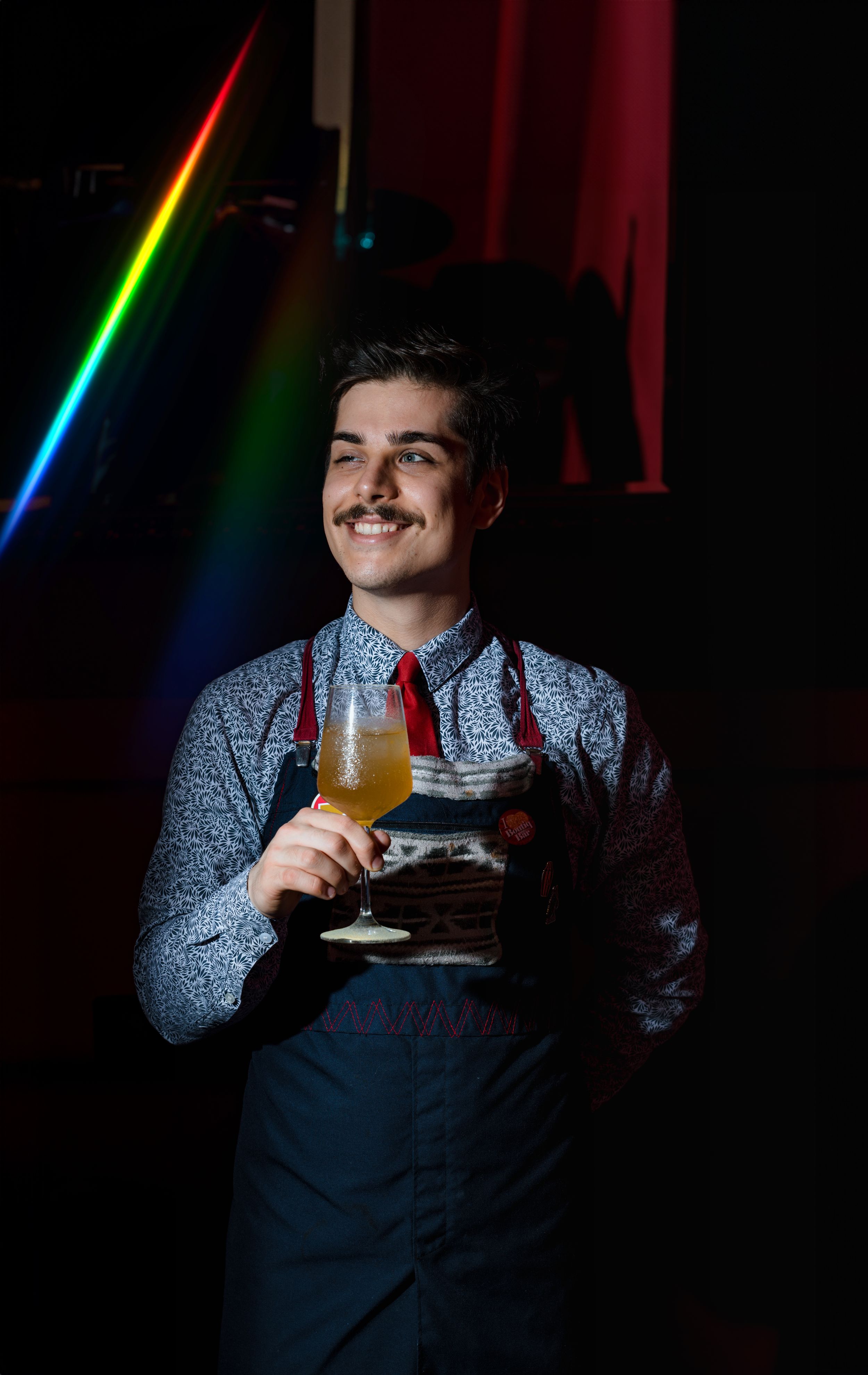 Bartender in apron holds cocktail with rainbow light effect in dark bar setting.