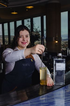 A bartender in an apron preparing drinks at a bar with a cityscape view.