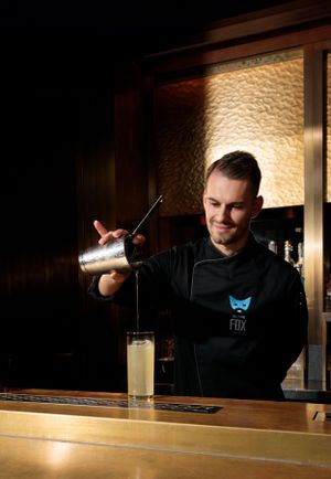 A bartender in black uniform pouring a cocktail from a shaker at a bar counter.