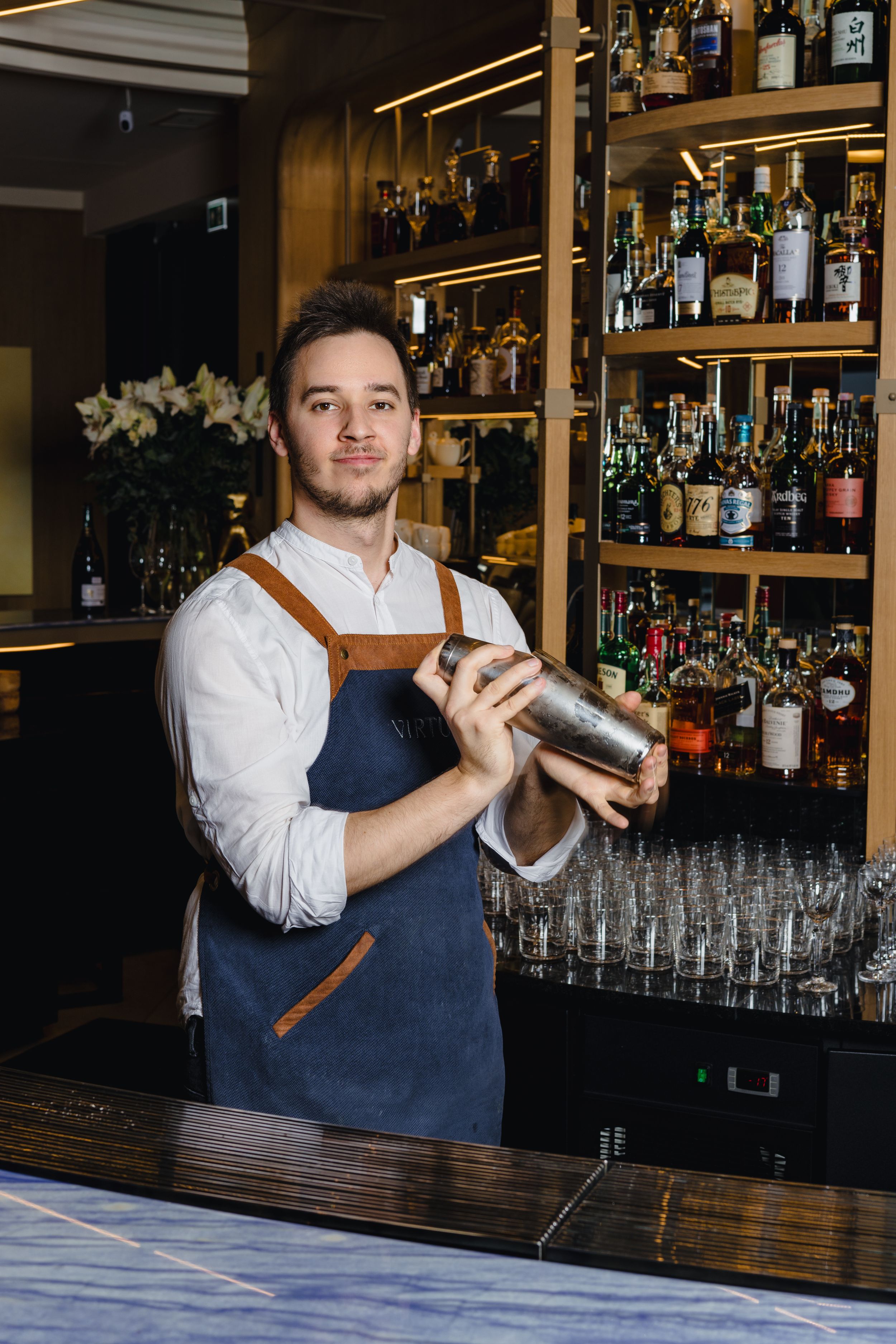 Professional bartender in apron preparing cocktails at well-stocked bar with extensive liquor selection.