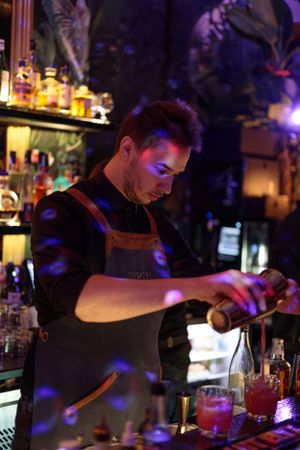A bartender in dark apron mixing drinks under purple ambient lighting.