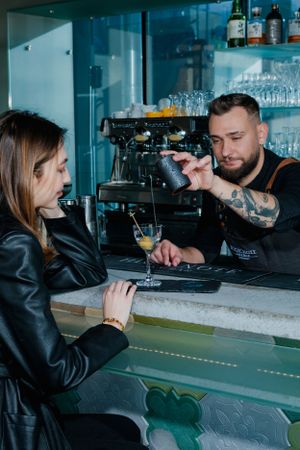 Bartender behind a bar counter interacts with a customer in a leather jacket during drink service.