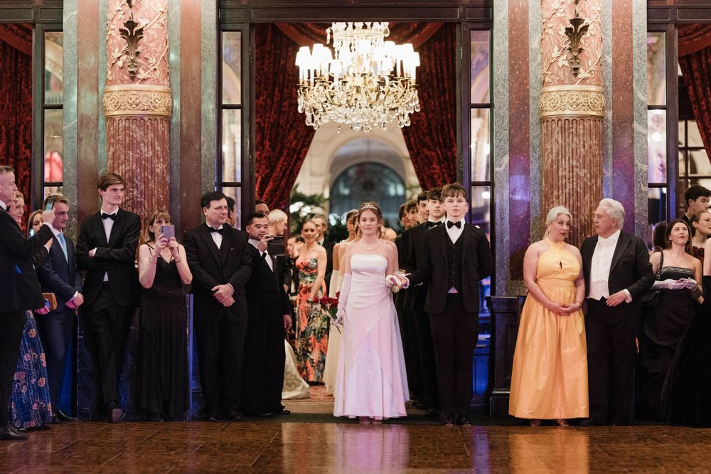 A formal ballroom celebration with attendees in elegant evening wear standing beneath a crystal chandelier.