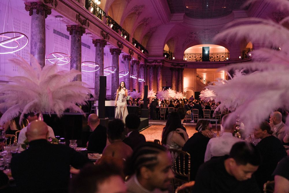 Elegant ballroom event with purple lighting, tall columns, and white feather decorations amid a seated audience.