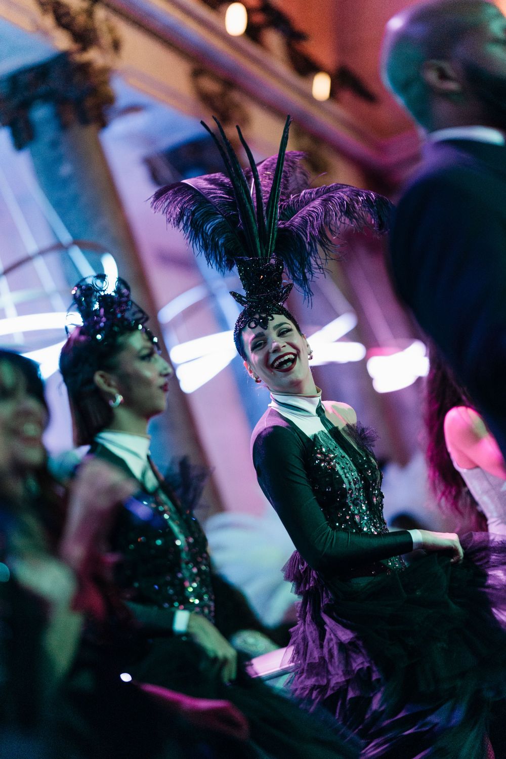 Performers in glamorous 1920s flapper style costumes with feather headdresses dance and smile during a cabaret show.