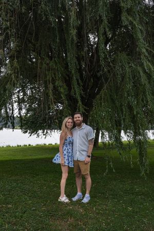 A romantic couple poses together under a drooping willow tree in a lush green park setting.