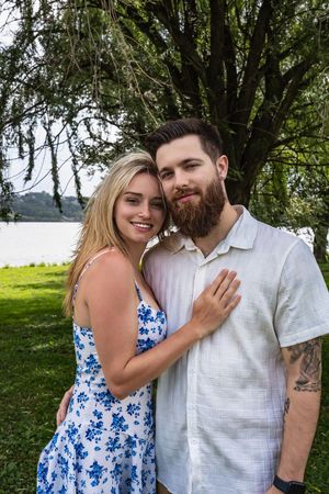 A couple poses together under a tree by a lake, with her wearing a blue floral dress and him in a white shirt.