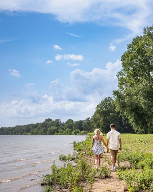 A couple walks along a scenic waterfront path with trees and blue sky on a sunny summer day.