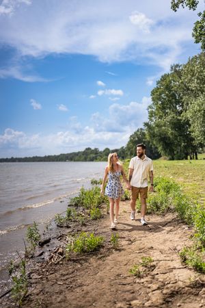 A couple walks along a scenic riverbank trail with blue skies and water views in the background.