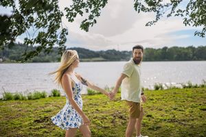 A couple walks hand in hand along a grassy waterfront area on a sunny day.