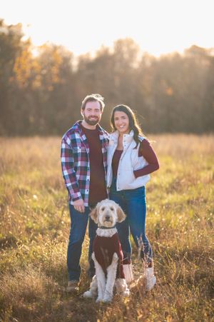 Couple poses with their goldendoodle dog in autumn field during golden hour.