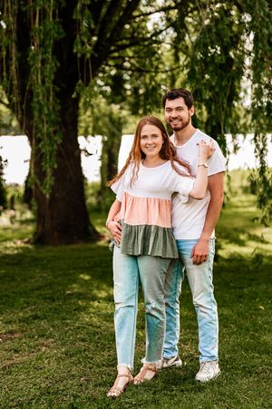 Casual couple in coordinated outfits standing together on green lawn with willow trees in background.