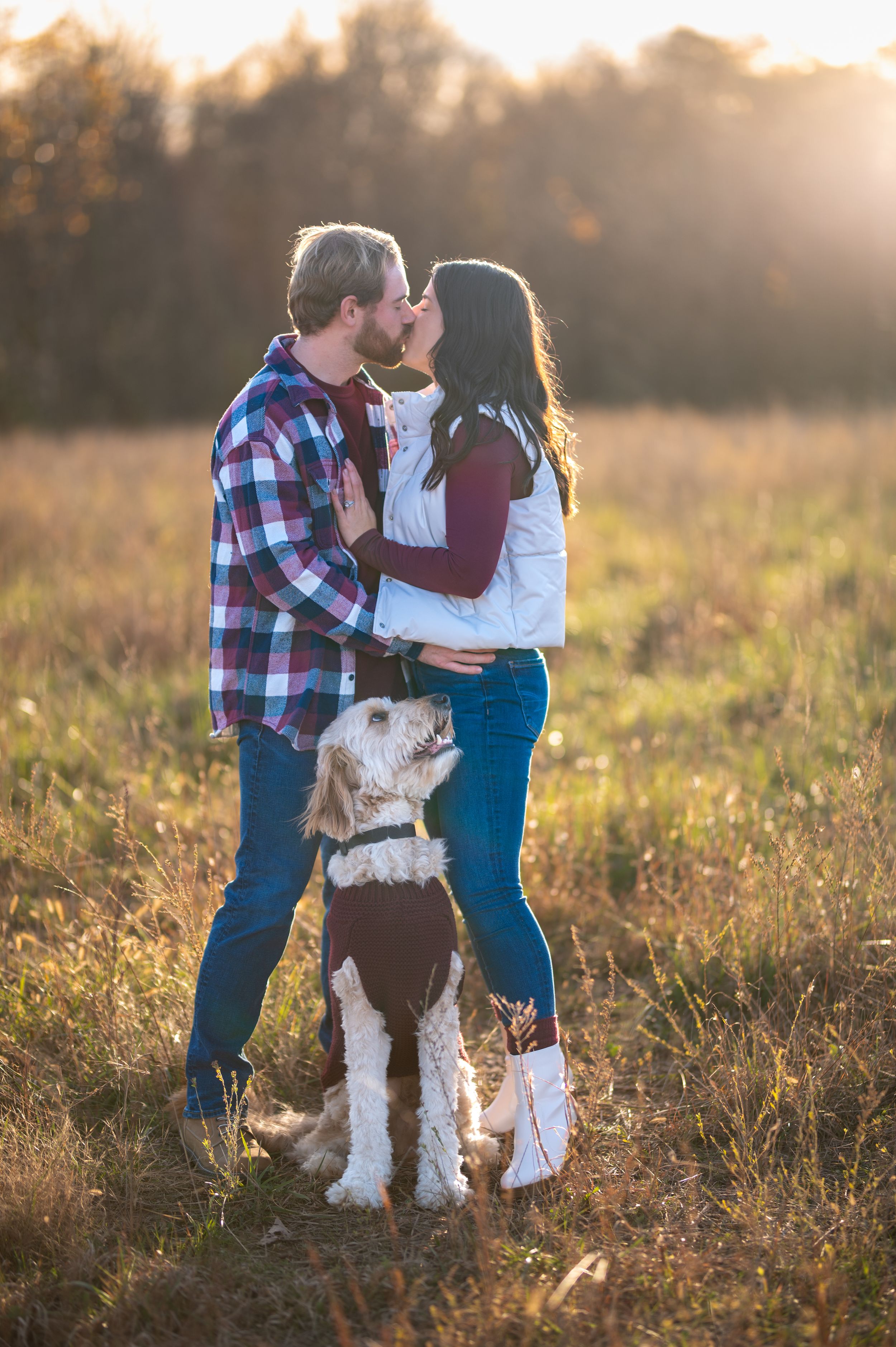 A couple shares a tender moment in a sunlit field with their white fluffy dog during a golden hour photoshoot.