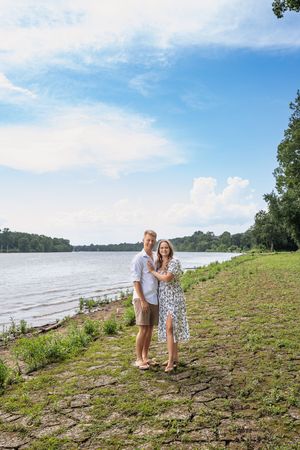 A couple stands together on a grassy hill overlooking a serene lake or river.