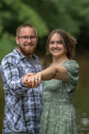 Engaged couple showing off ring while posing together by a creek.