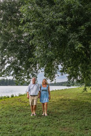 A couple stands beneath a large shady tree on a grassy lawn overlooking a serene lake.