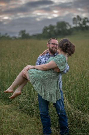 Man carrying woman in green dress across field during sunset engagement photo session.