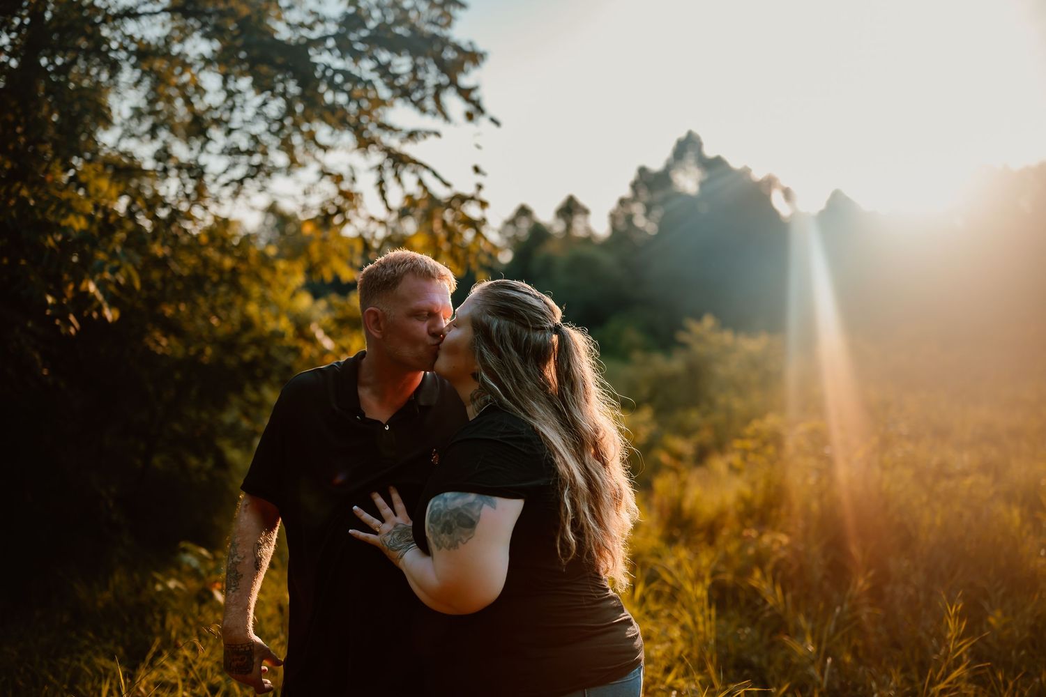 A couple embraces in a golden sunlit field during sunset with trees in the background.