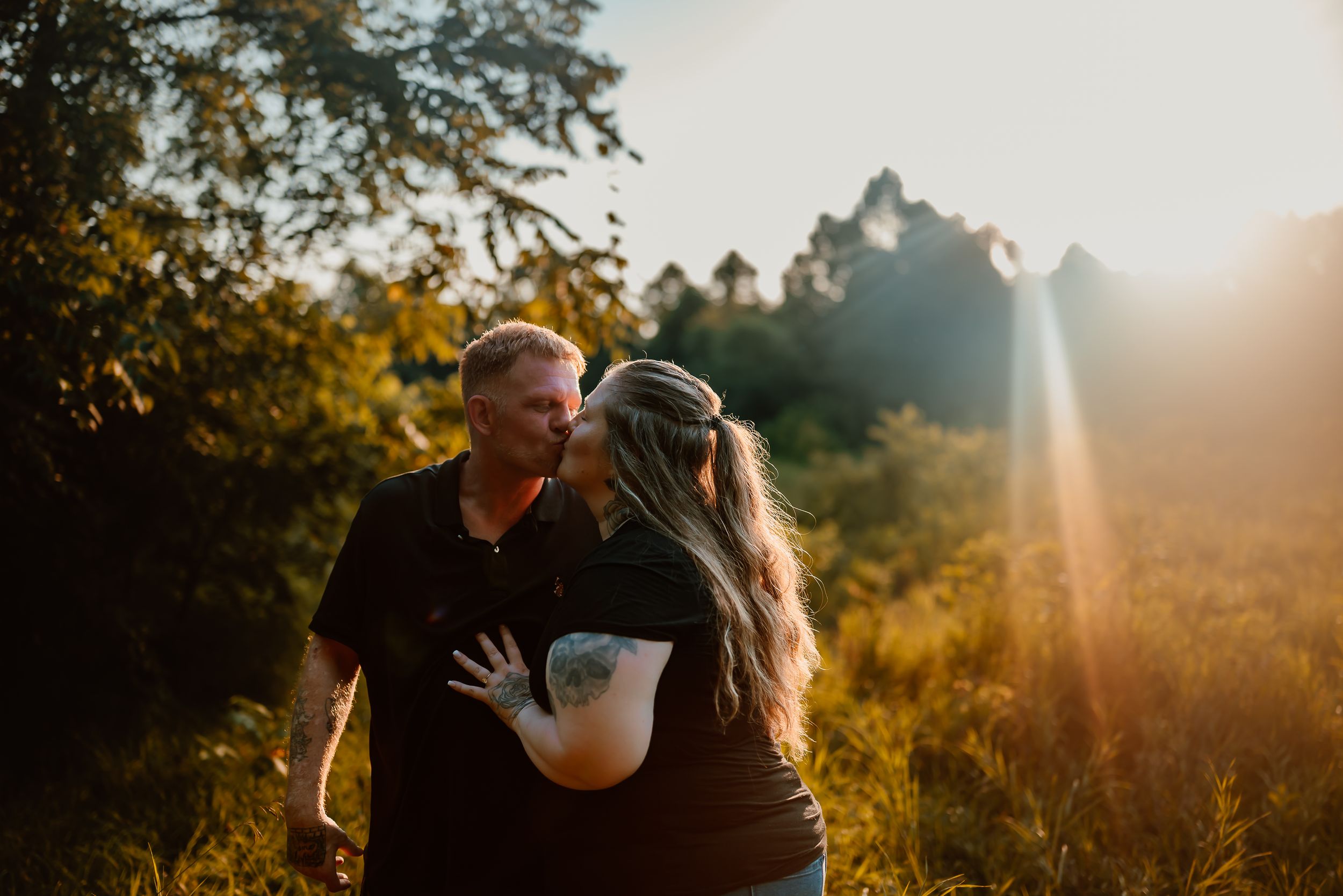 A couple embraces in a golden sunlit field during sunset with trees in the background.