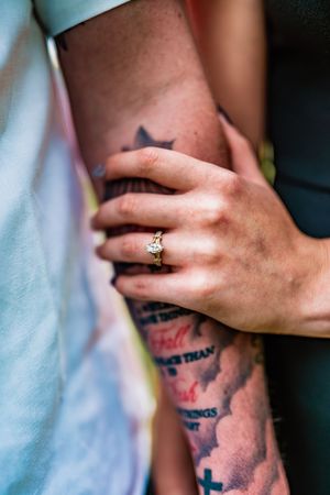 A hand wearing an engagement ring gently rests on a tattooed arm with dark ink designs visible.