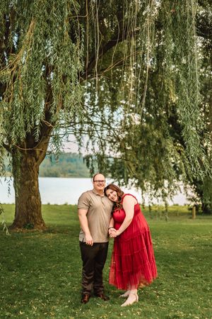 A couple enjoys a romantic moment under drooping willow trees by a serene lakeside setting.