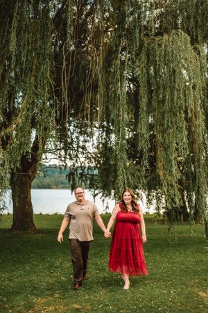 A couple walks together underneath drooping willow tree branches on a lush green lawn near a lake.