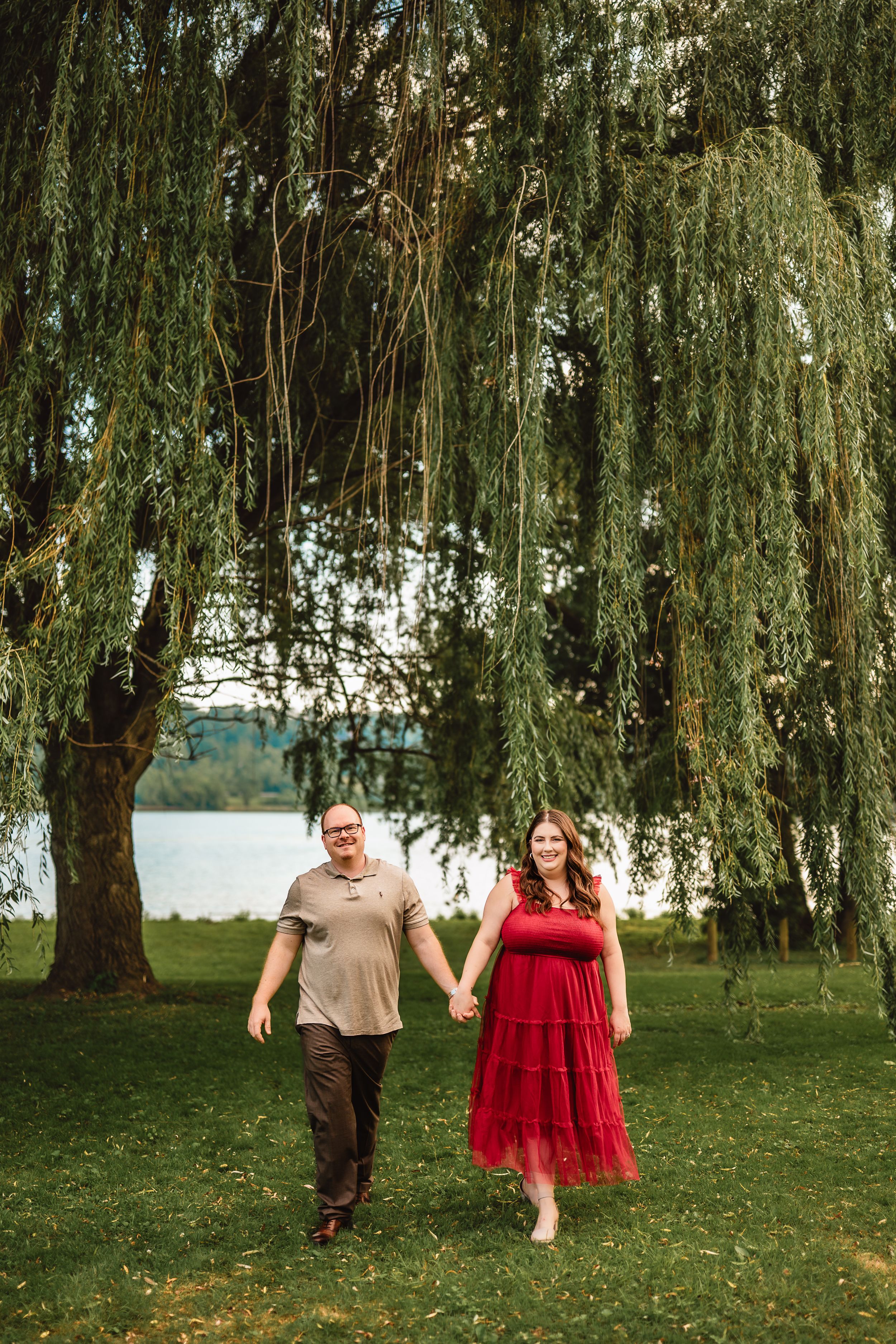 A couple walks together underneath drooping willow tree branches on a lush green lawn near a lake.