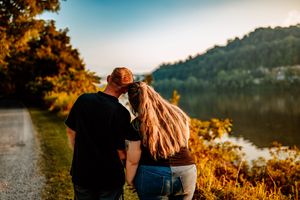 A couple embraces against a golden sunset backdrop near a lake with mountains in the background.