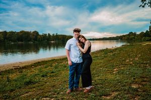 A couple shares a romantic moment on a grassy hillside overlooking a serene lake during sunset.