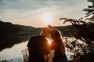 Silhouettes of a couple embrace against a dramatic sunset by a serene lake as trees frame the romantic moment.