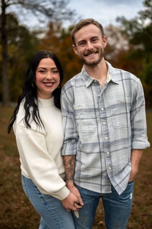 A couple in casual fall attire smiles together outdoors with autumn foliage in the background.