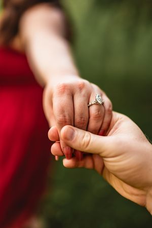 Close-up series showing a hand passing a delicate diamond engagement ring to another hand against a blurred green background.