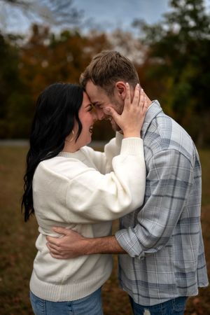 A romantic couple embraces outdoors during autumn, wearing casual attire against a backdrop of fall foliage.